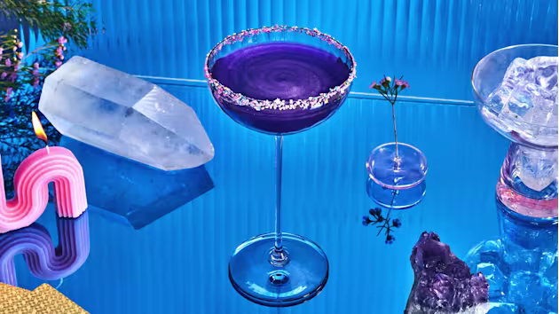 A purple cocktail in a coupe glass with a decorative rim sits on a reflective blue surface, surrounded by crystals, ice, a pink candle, and a small flower in a vase.