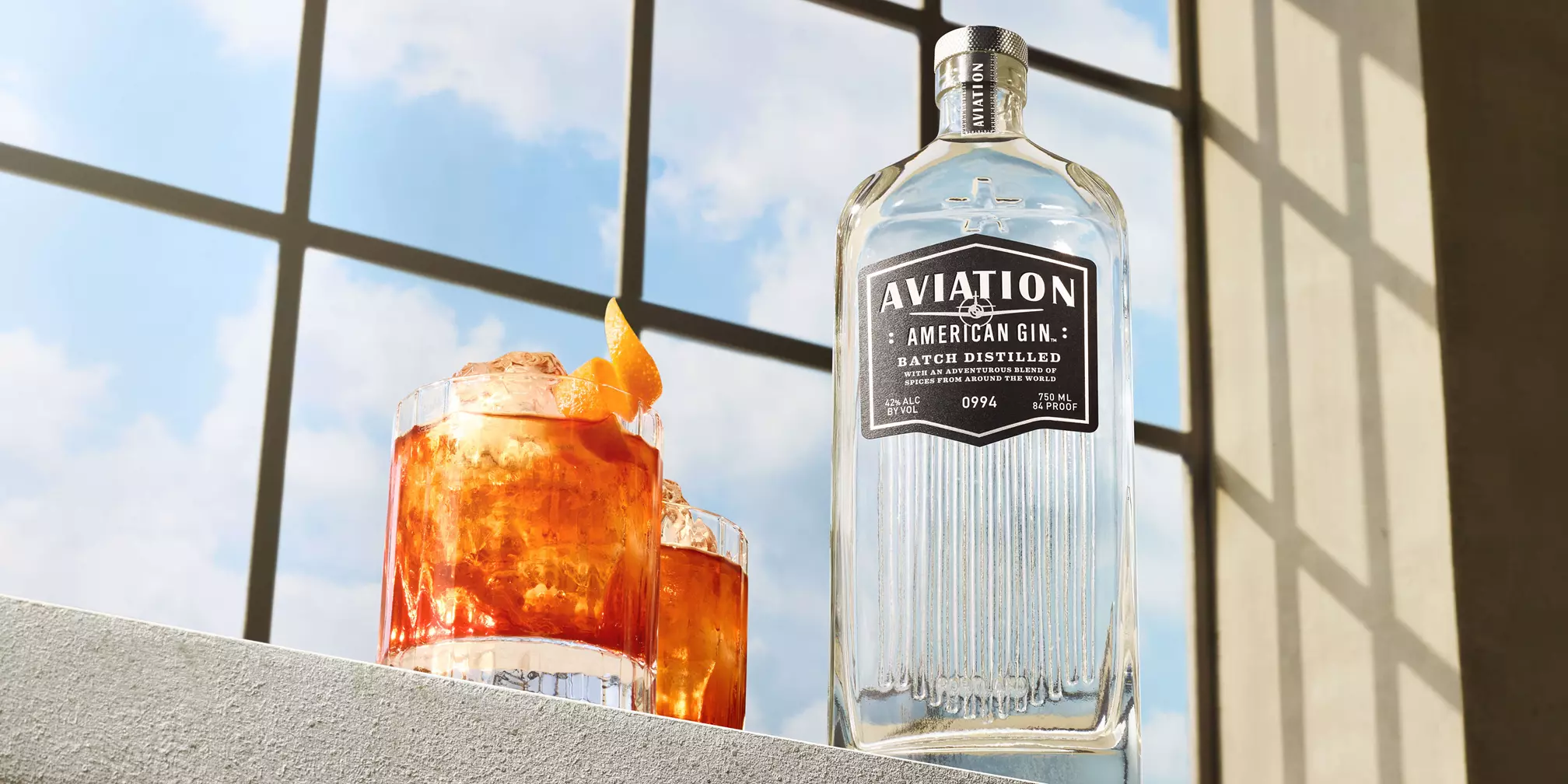 A bottle of Aviation American Gin stands next to two cocktails with orange garnishes, set on a ledge in front of a sunlit window.