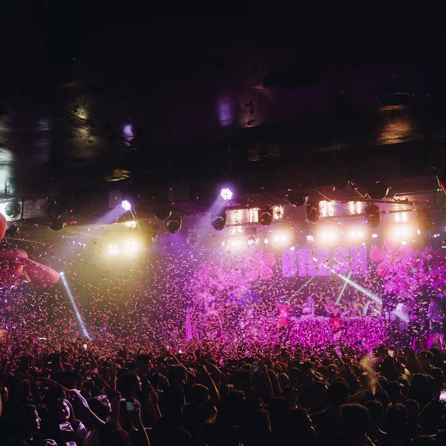 La foto muestra una escena de concierto en La Riviera, con una gran multitud de personas. El escenario está iluminado con luces y hay efectos visuales como haces de luz y lo que parece ser confeti en el aire.