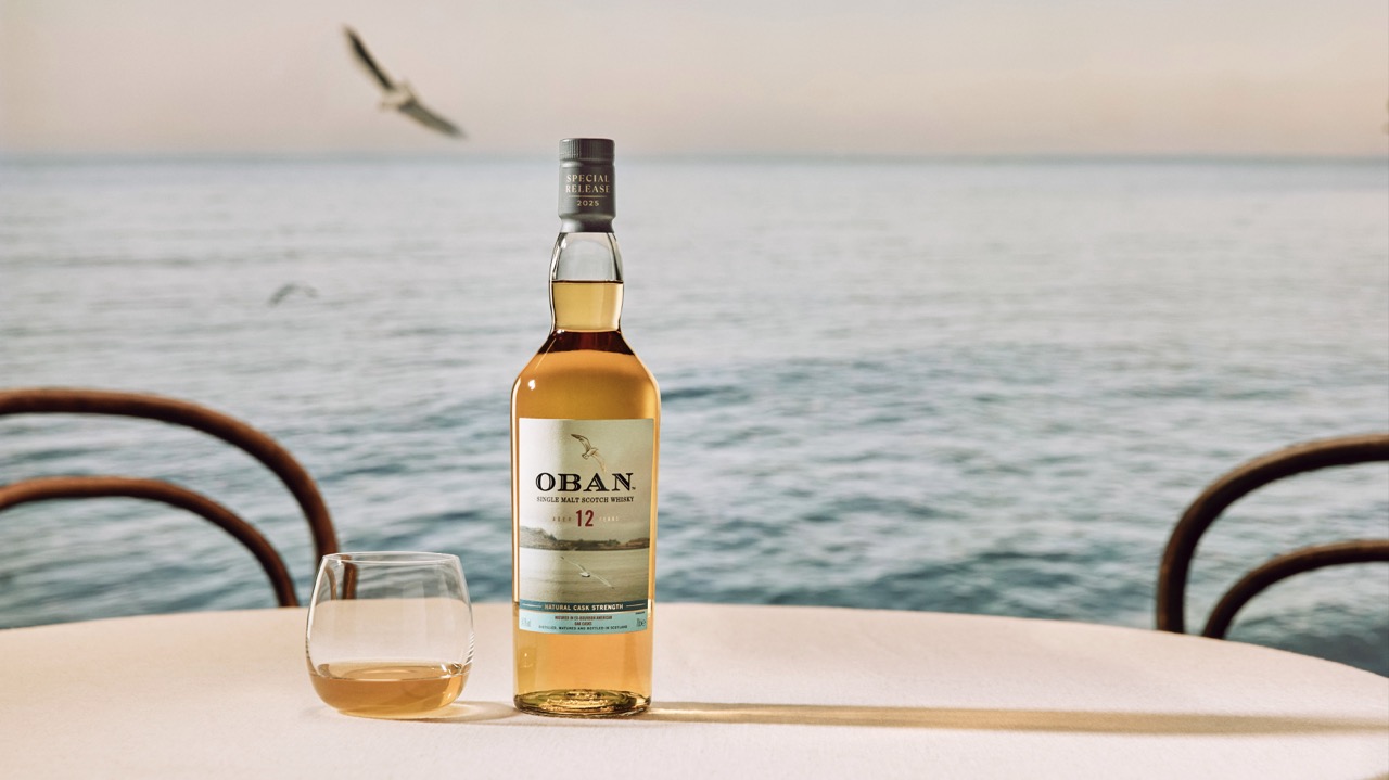 A bottle of Oban 12-year-old whisky and a glass with whisky sit on a round table by the sea, with a bird flying over the water in the background.