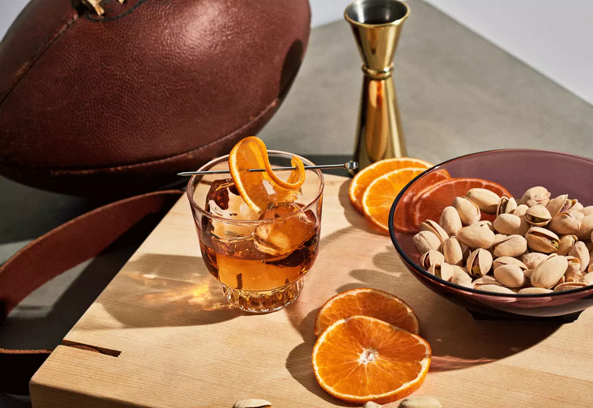 A glass of cocktail with ice and orange peel, a bowl of pistachios, orange slices, a jigger, and a football on a wooden board.
