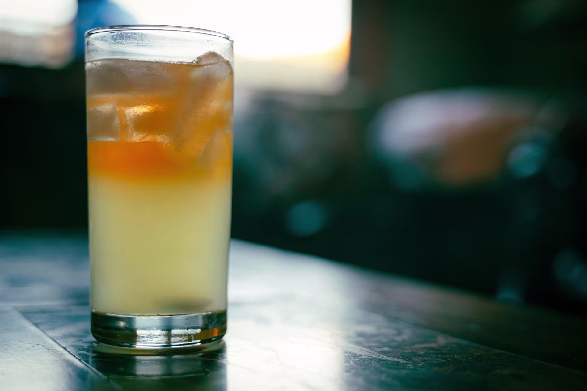 A glass filled with a layered iced drink, showing a gradient from pale yellow at the bottom to amber at the top, placed on a table.