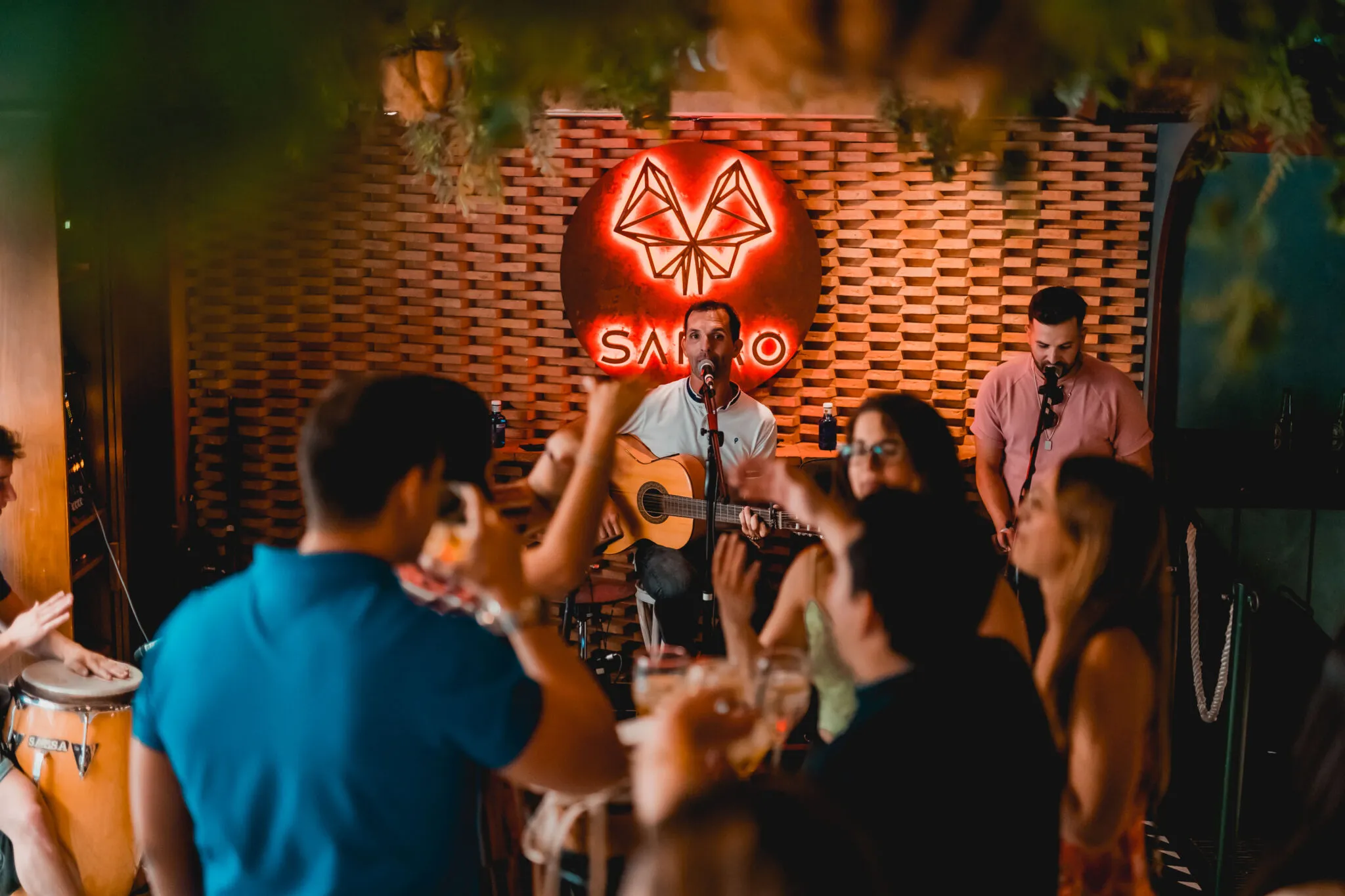La imagen muestra un ambiente cálido en Sarao, con un músico tocando guitarra y cantando frente a una pared iluminada. El público disfruta animado.