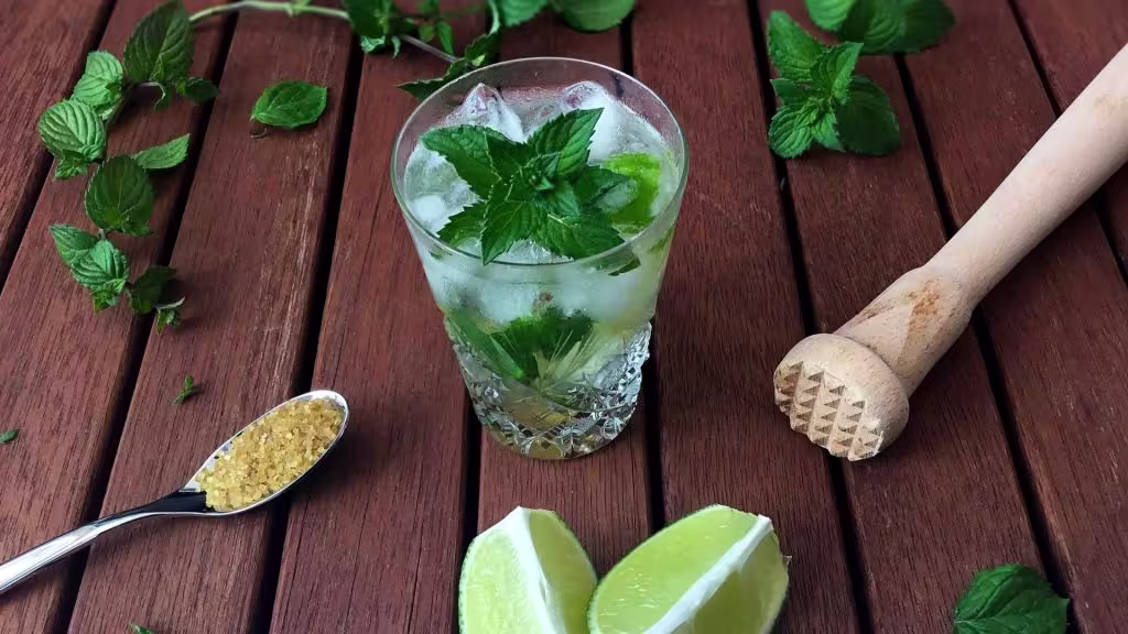A glass of mojito cocktail with mint leaves and ice on a wooden table, surrounded by fresh mint, a muddler, lime wedges, and a spoonful of brown sugar.