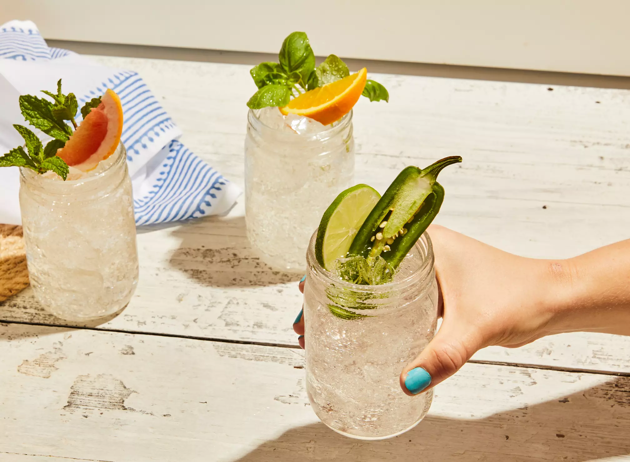 Three mason jars with crushed ice and garnishes; one with jalapeño and lime, one with mint and grapefruit, and one with basil and orange. A hand holds the jar with jalapeño and lime.