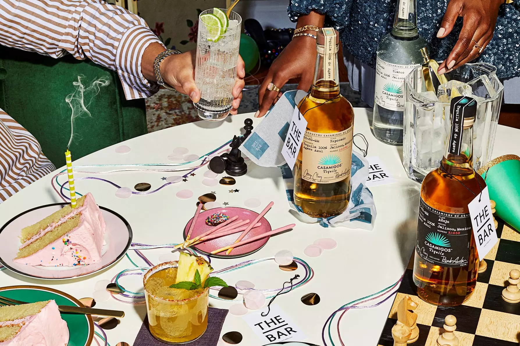 A table with cocktails, bottles of liquor, a slice of cake with a lit candle, party decorations, a chessboard, and two people’s hands preparing drinks.
