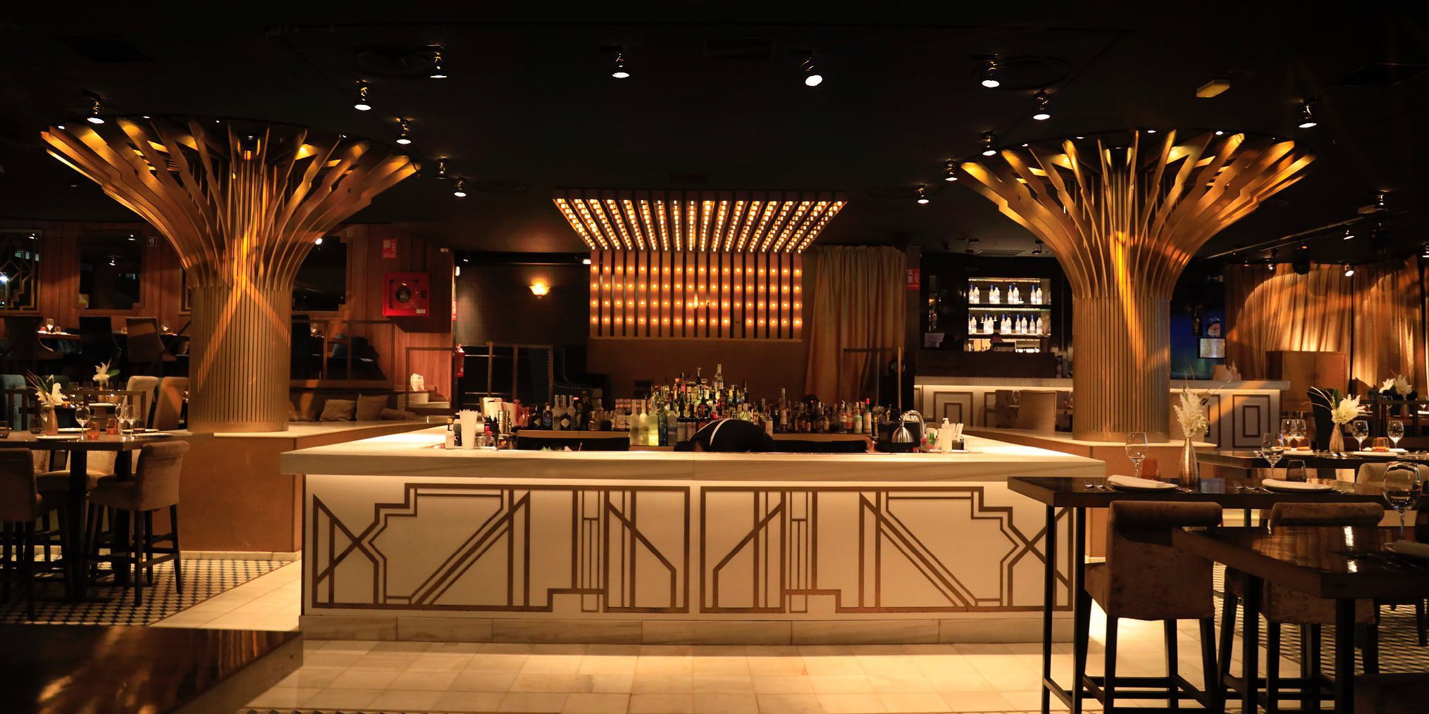 La imagen muestra el interior de Gatsby, un lugar elegante y sofisticado con columnas doradas de diseño artístico y una barra central bien surtida. La iluminación cálida y el mobiliario cuidado crean un ambiente de lujo y exclusividad.