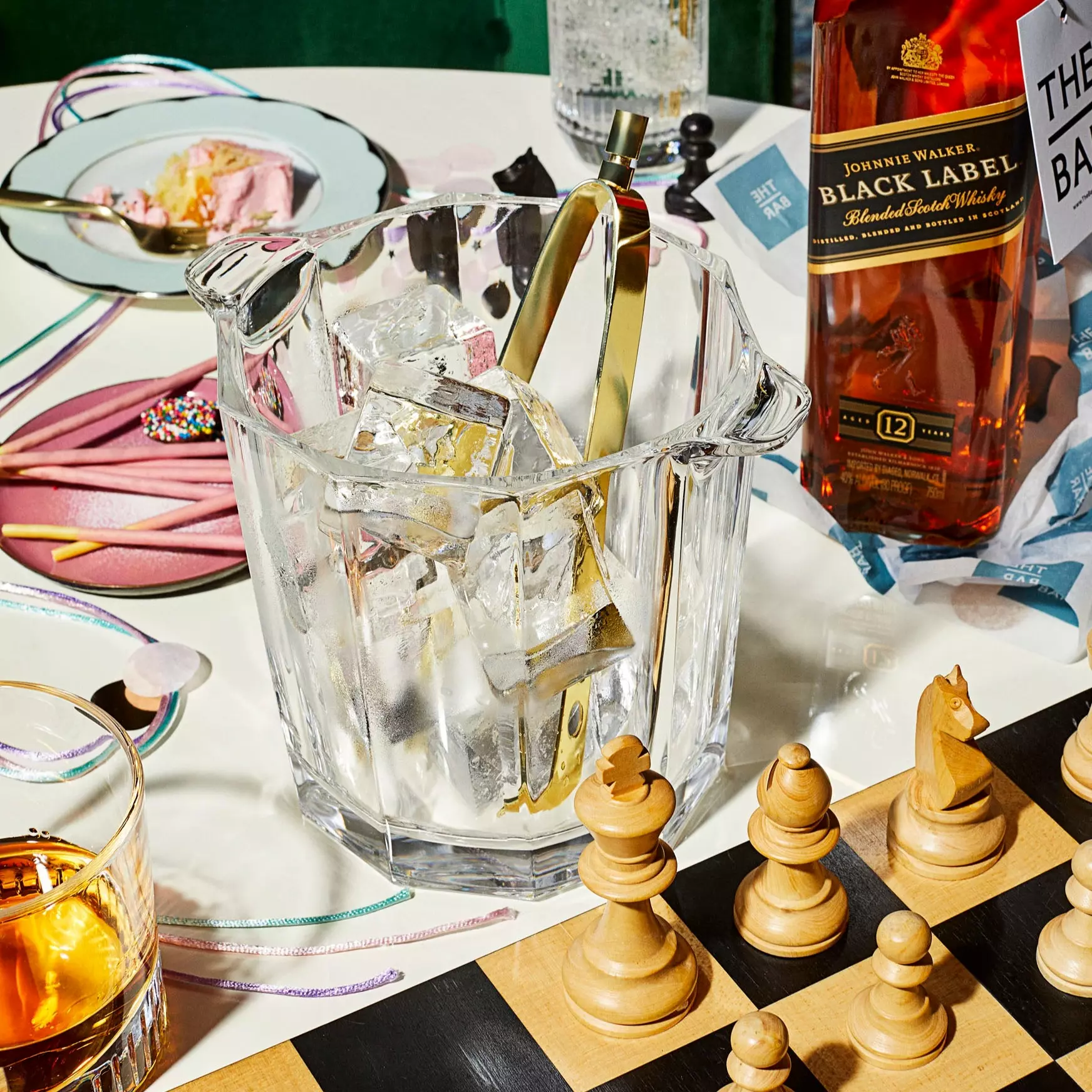 Glass ice bucket with tongs sits on a table next to a bottle of whiskey, a glass of liquor, a chessboard, and party remnants such as ribbons, cake, and confetti.