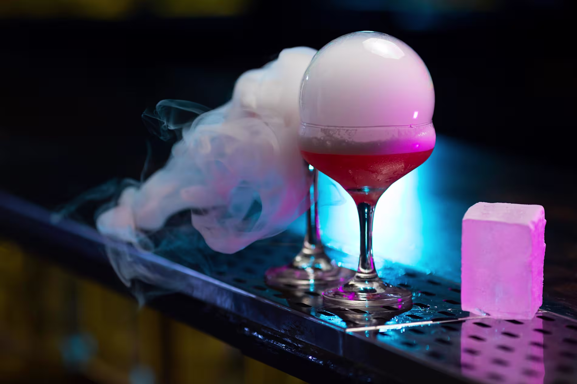 Two cocktail glasses with red drinks, topped with a smoke-filled bubble, next to a smoking chunk of ice on a bar counter.