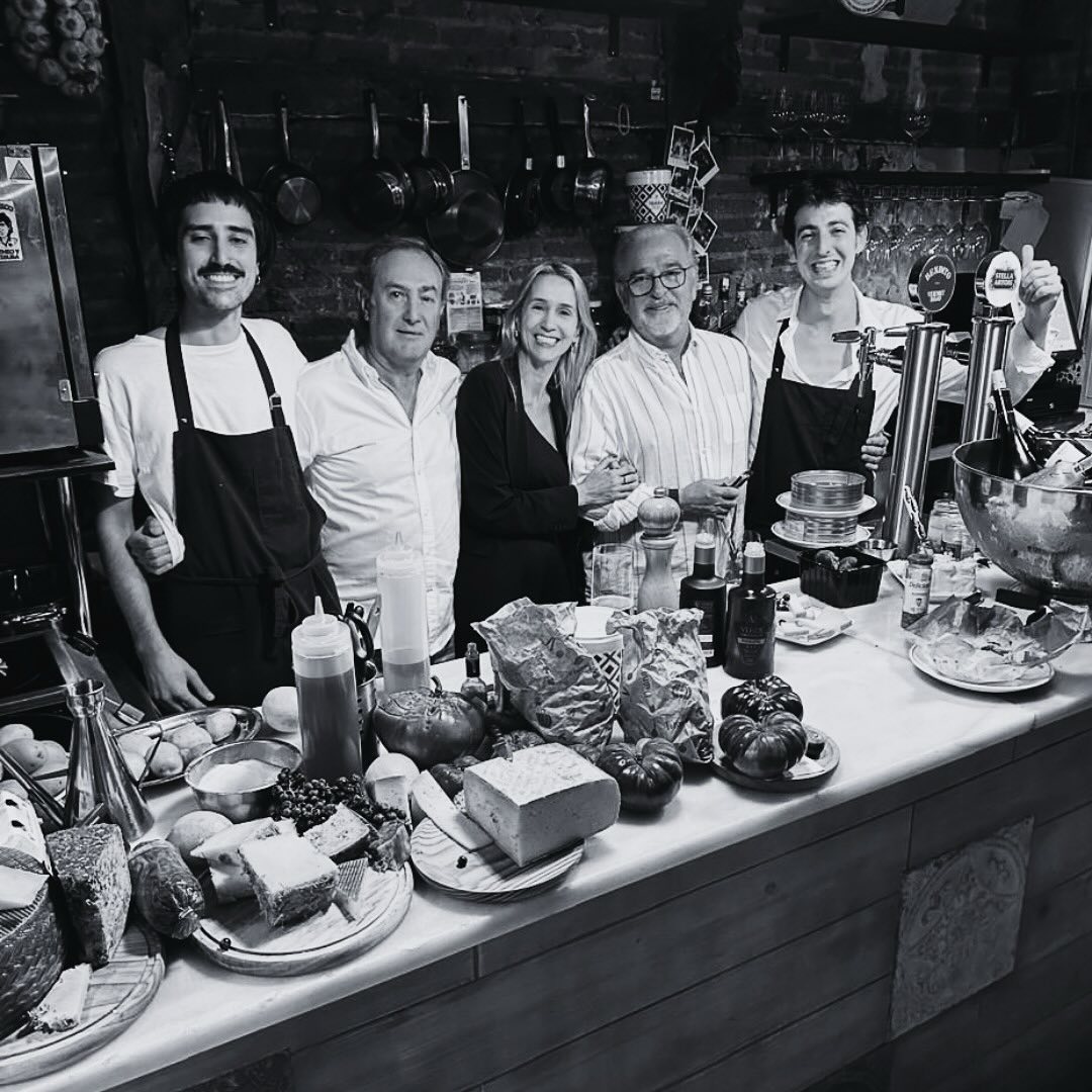 La foto muestra una imagen en blanco y negro de cinco personas detrás de un mostrador lleno de varios alimentos y utensilios de cocina.