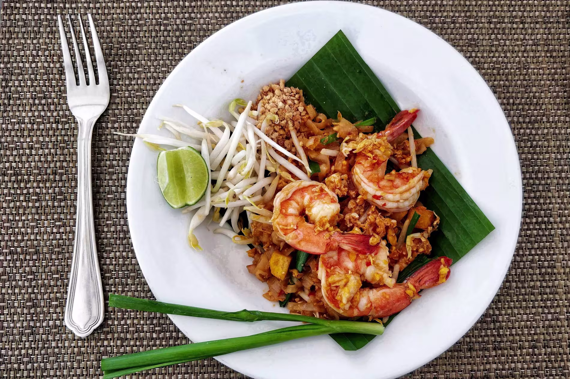 A plate of shrimp pad thai with bean sprouts, lime, chopped peanuts, and green onions on a banana leaf, placed next to a fork on a woven placemat.