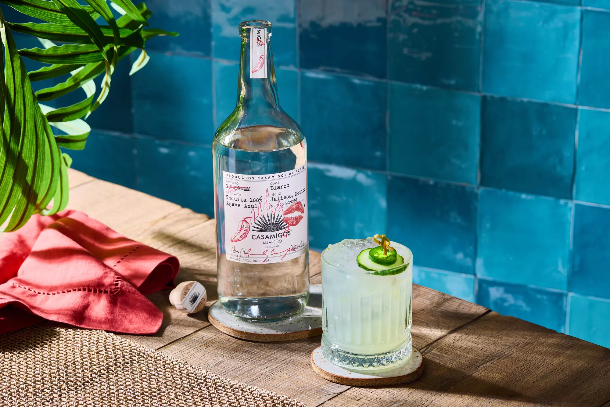 A bottle of Casamigos tequila and a glass of cocktail garnished with cucumber slices sit on a wooden table next to a red napkin and a blue tiled wall.
