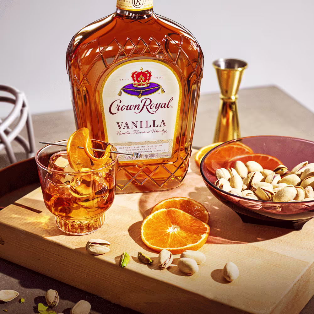 A bottle of Crown Royal Vanilla whiskey, a glass with whiskey and orange slice, a bowl of pistachios, and orange slices on a wooden board.