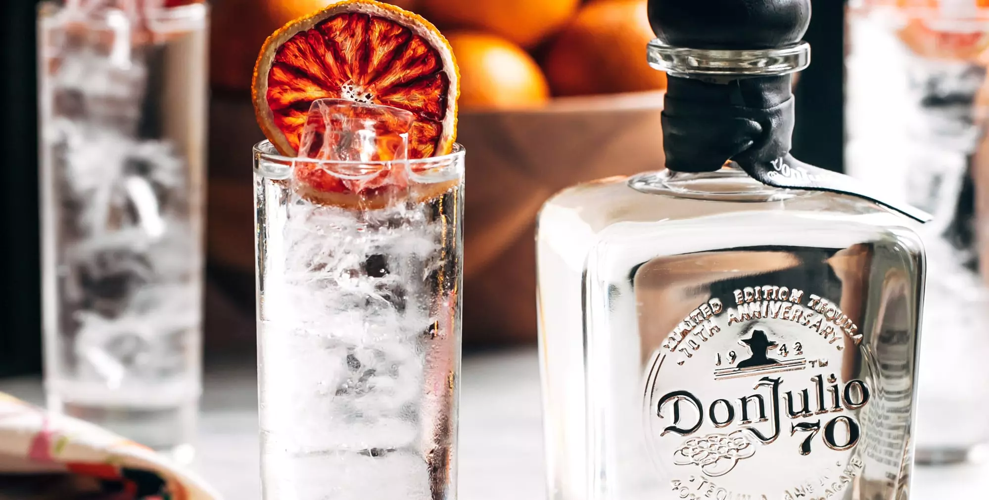 A glass filled with ice and a dried citrus slice sits next to a bottle of Don Julio 70 tequila; oranges are visible in a basket in the background.