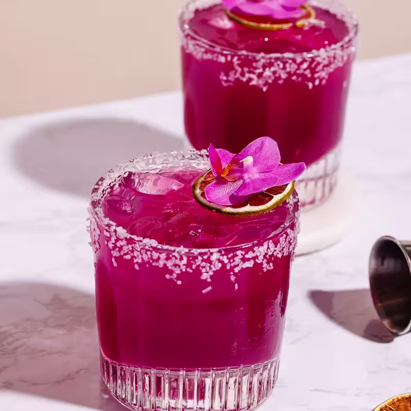 Two vibrant purple cocktails with salted rims, ice, and garnished with a slice of lime and a purple orchid flower on a white marble surface.