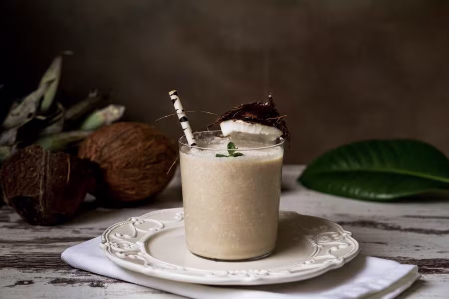 A glass of creamy coconut smoothie with a straw, garnished with a coconut piece and mint, sits on a white plate and napkin, with coconuts and a green leaf in the background.