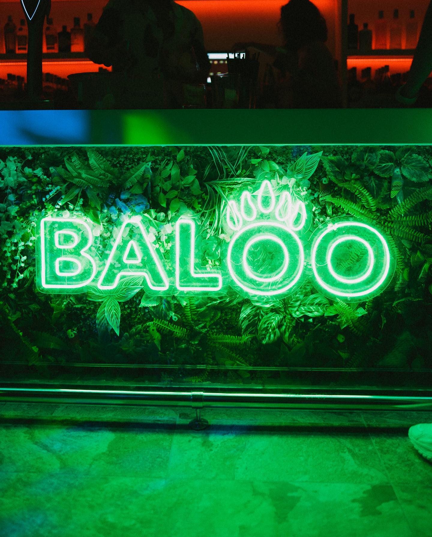 Letrero de neón verde de Baloo rodeado de plantas, creando un ambiente fresco y moderno en el interior del bar.