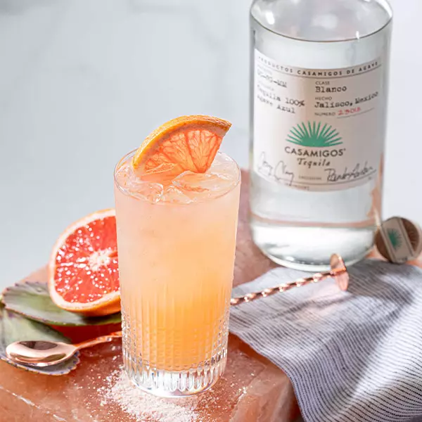 A tall glass of pink cocktail with ice and a grapefruit slice garnish sits beside a bottle of Casamigos Tequila Blanco and a halved grapefruit.