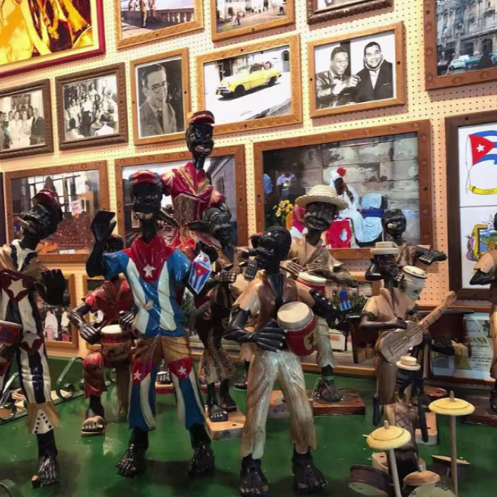 Colección de figuritas coloridas, representando músicos, ya que están sosteniendo varios instrumentos. Las figuras parecen estar en una habitación con paredes cubiertas de fotografías y obras de arte enmarcadas. Todas las figuritas tienen temática Cubana.