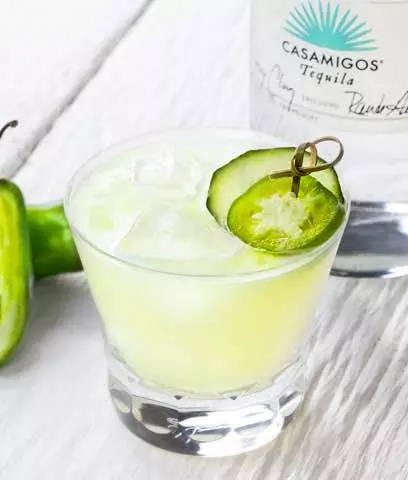 A glass of light green cocktail with ice, garnished with cucumber and jalapeño slices, sits beside a bottle of Casamigos Tequila on a white surface.