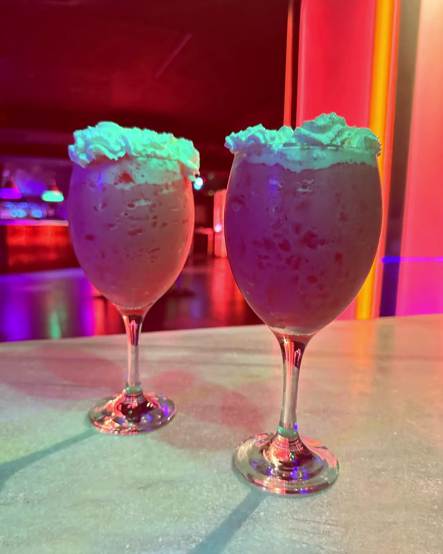 Bisu con dos cócteles cremosos y coloridas luces de fondo, creando una atmósfera festiva y divertida para disfrutar la noche.