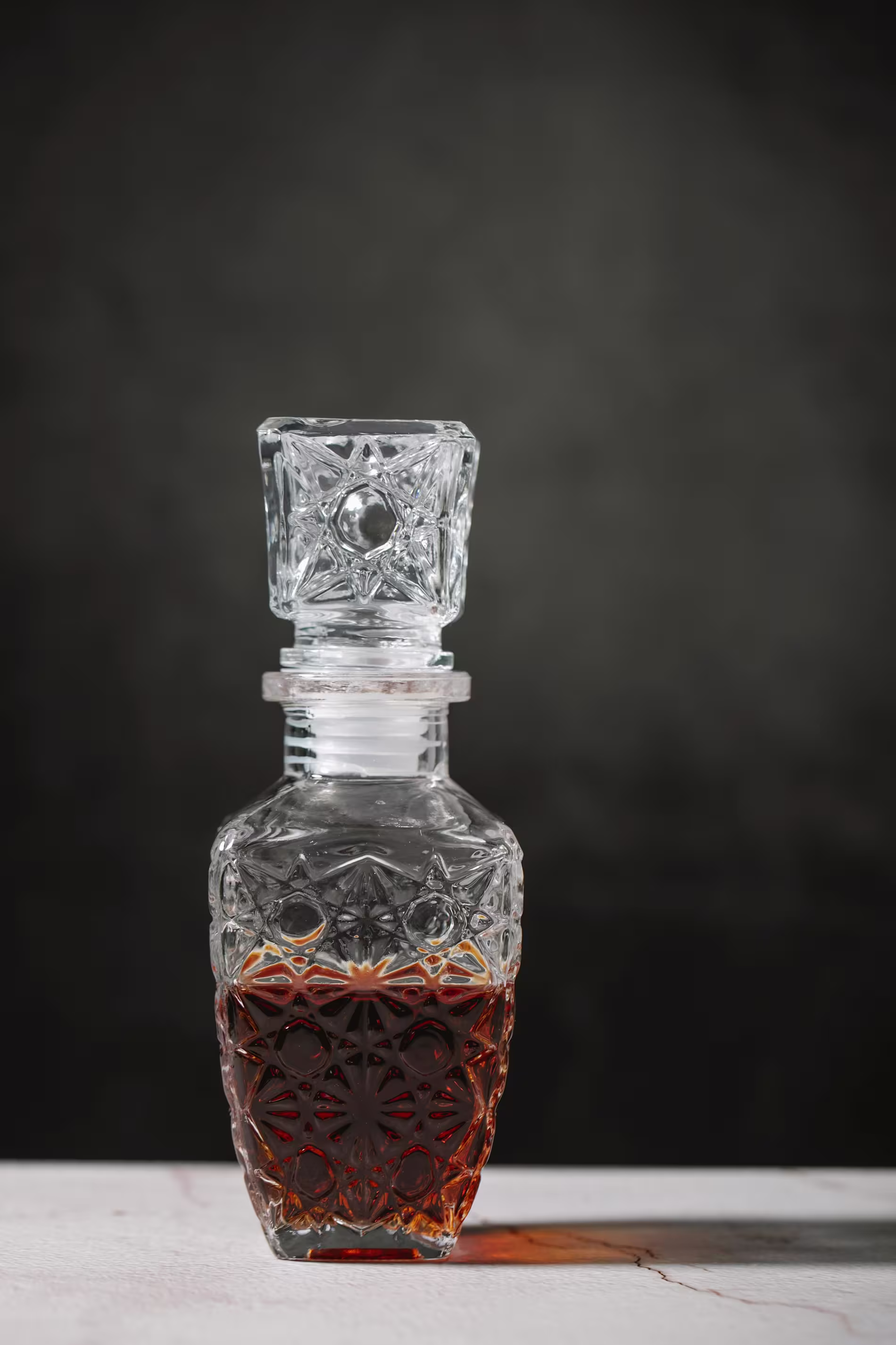 A glass decanter with an ornate pattern filled with a dark amber liquid, placed on a light surface against a dark background.