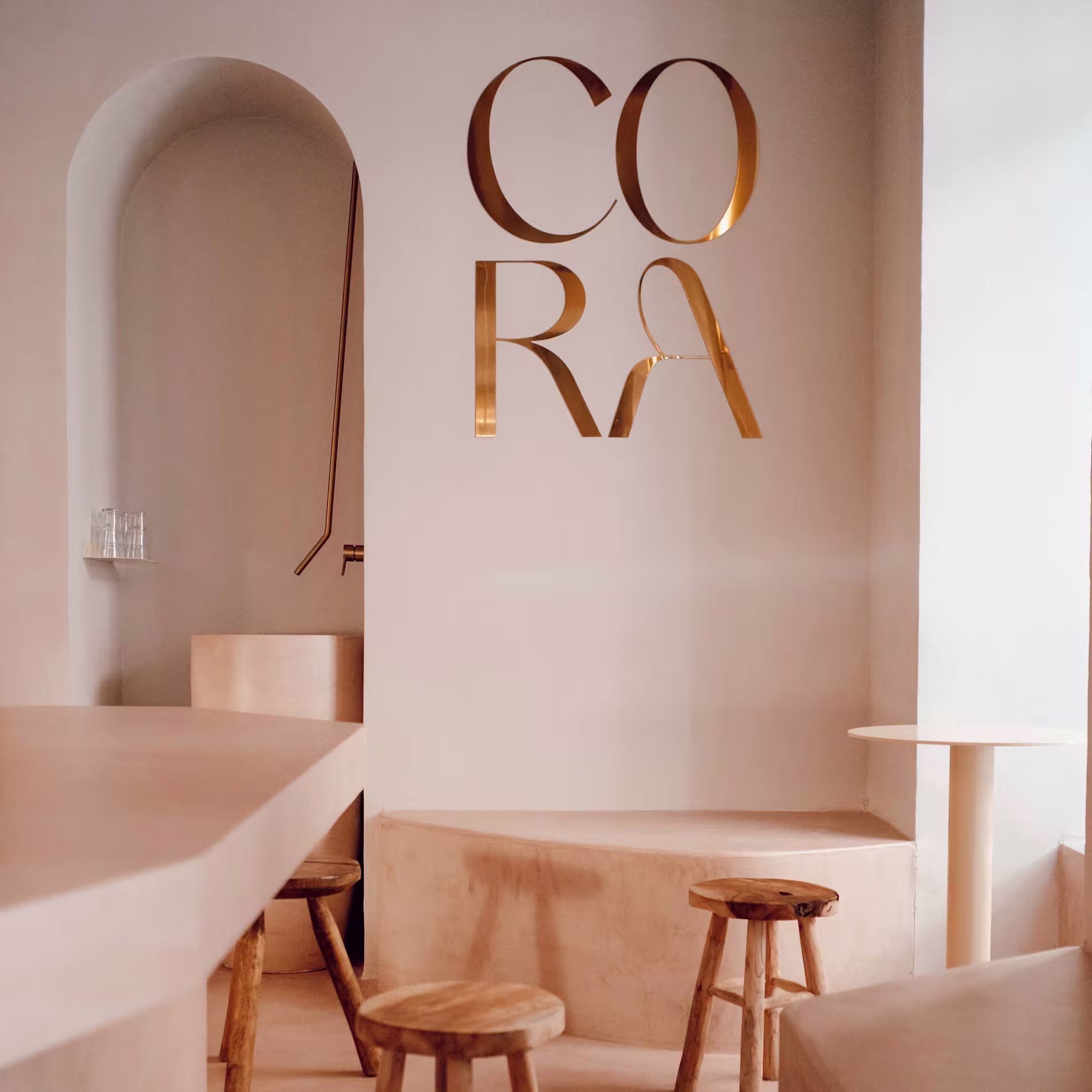 Interior minimalista de Cora con tonos tierra, taburetes de madera, formas curvas y el nombre del local en letras doradas sobre pared blanca.