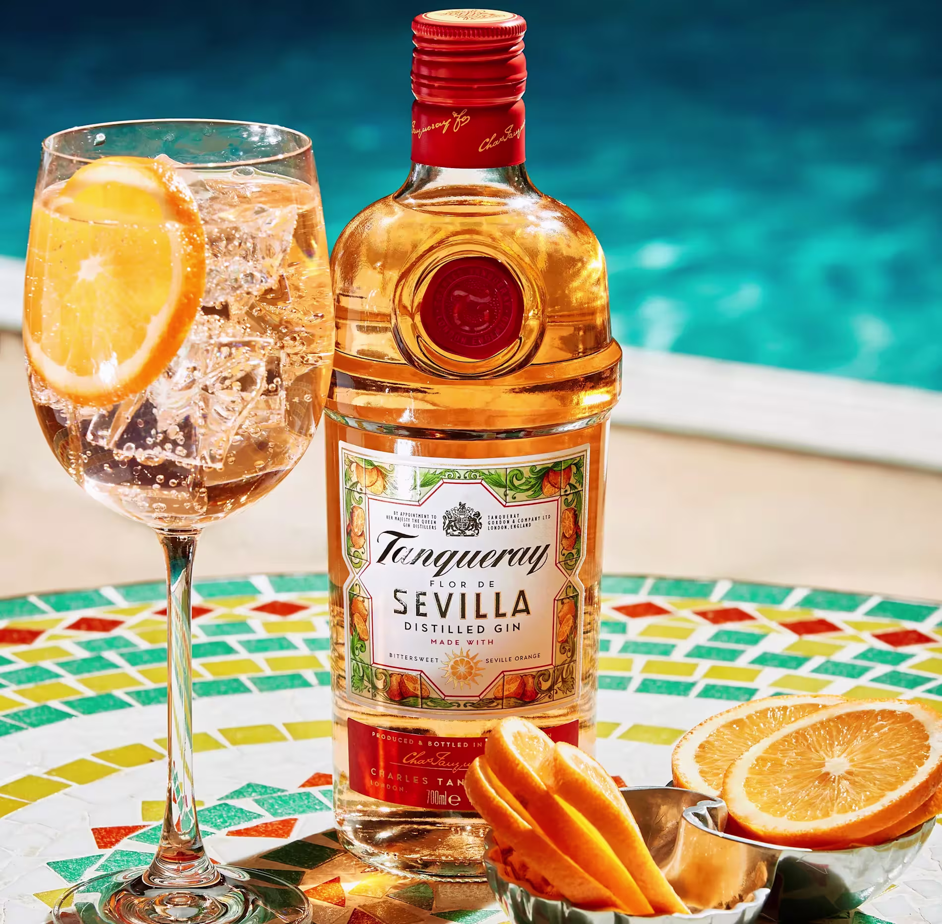 A bottle of Tanqueray Flor de Sevilla gin stands next to a cocktail with an orange slice, a small bowl of orange slices, all placed on a colorful mosaic table by a pool.