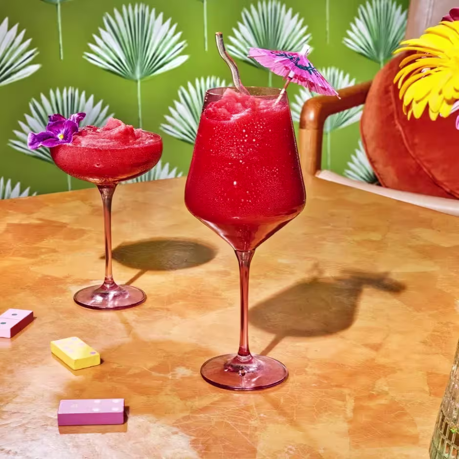 Two red frozen cocktails in stemmed glasses sit on a table; one has a pink umbrella and straw, the other an orchid. Colorful candies and flowers are also visible.