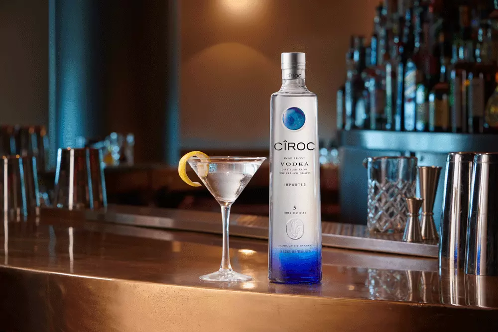 A bottle of Ciroc vodka and a martini glass with a lemon twist sit on a bar counter, with barware and bottles in the background.