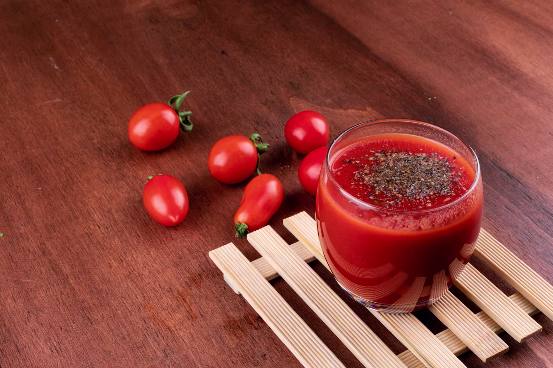 A glass of tomato juice topped with ground black pepper sits on a wooden mat, with several whole cherry tomatoes scattered on a wooden table.