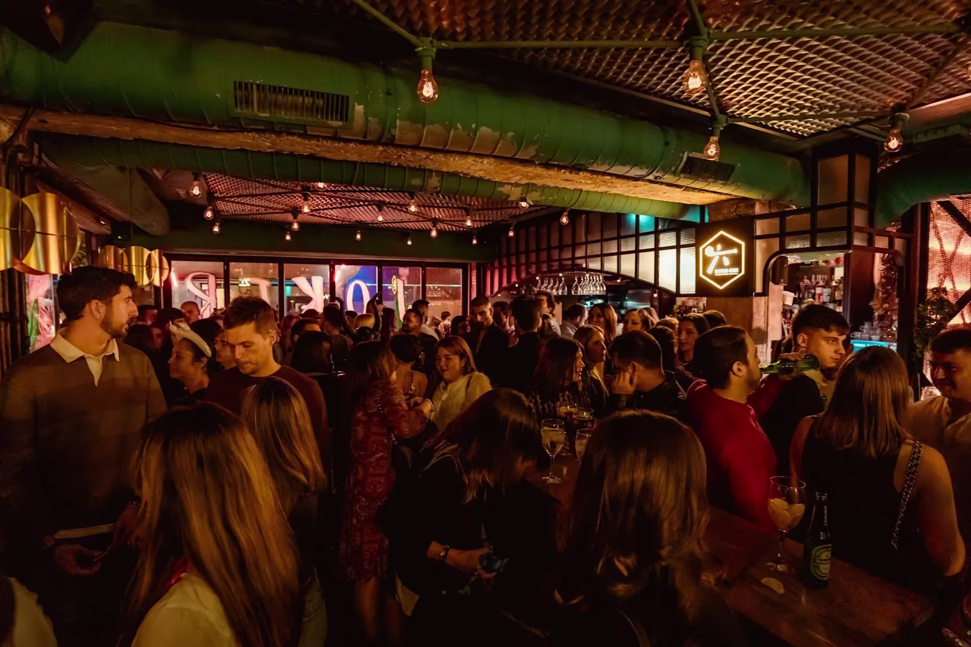 La imagen muestra un ambiente animado en Sarao, un bar o club con luces tenues y una multitud disfrutando de cócteles y música en un espacio moderno.