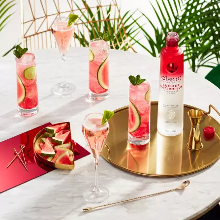 A round table with glasses of pink cocktails garnished with lime and mint, a bottle of Ciroc Summer Watermelon, watermelon slices, and cocktail tools.