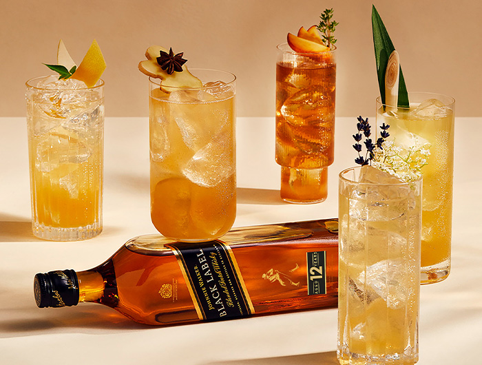 A bottle of Johnnie Walker Black Label whisky lying on a table surrounded by six cocktails garnished with fruit and herbs.