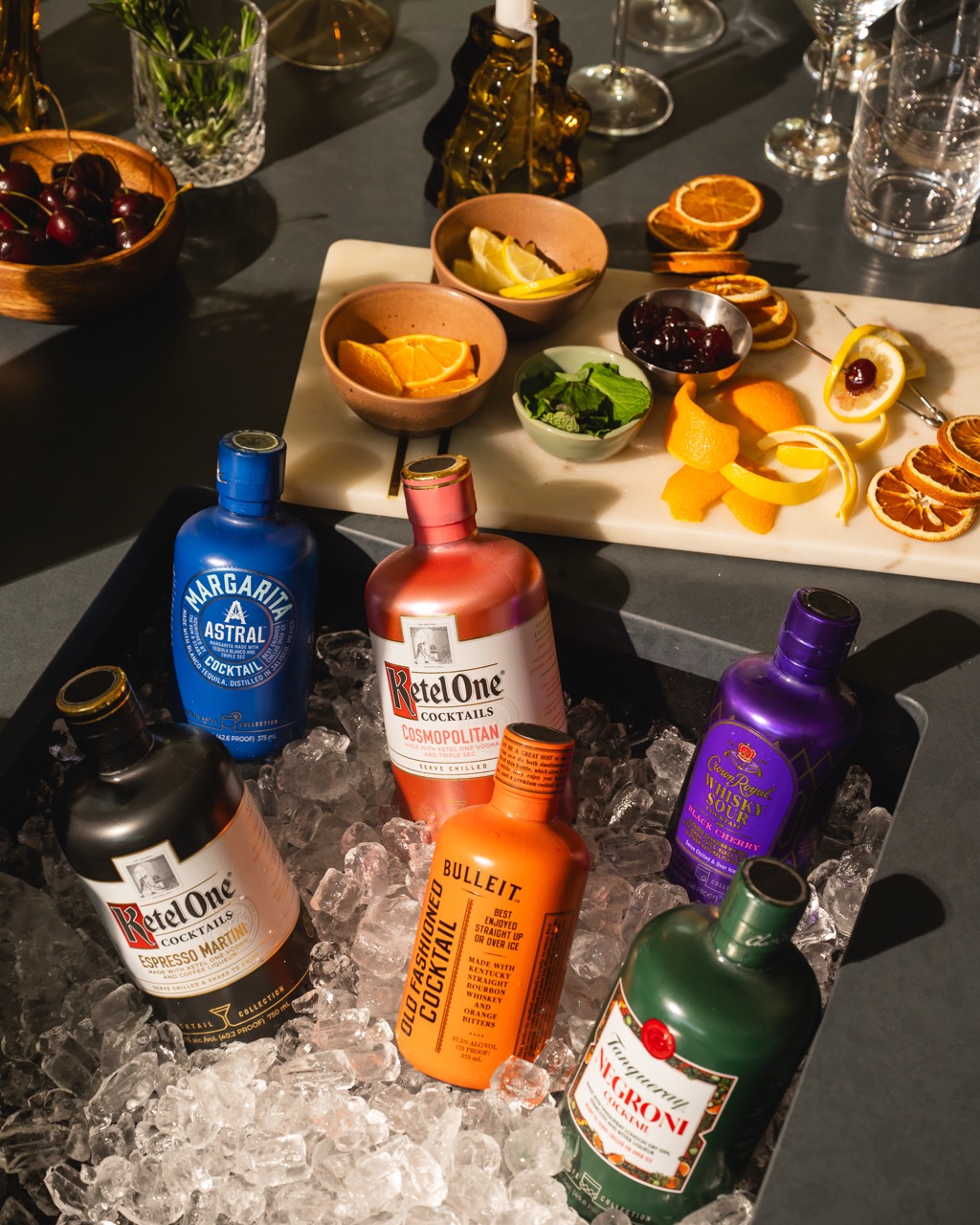 Five bottled cocktails rest on ice in a tray, surrounded by cocktail garnishes, fruits, and glassware on a table.