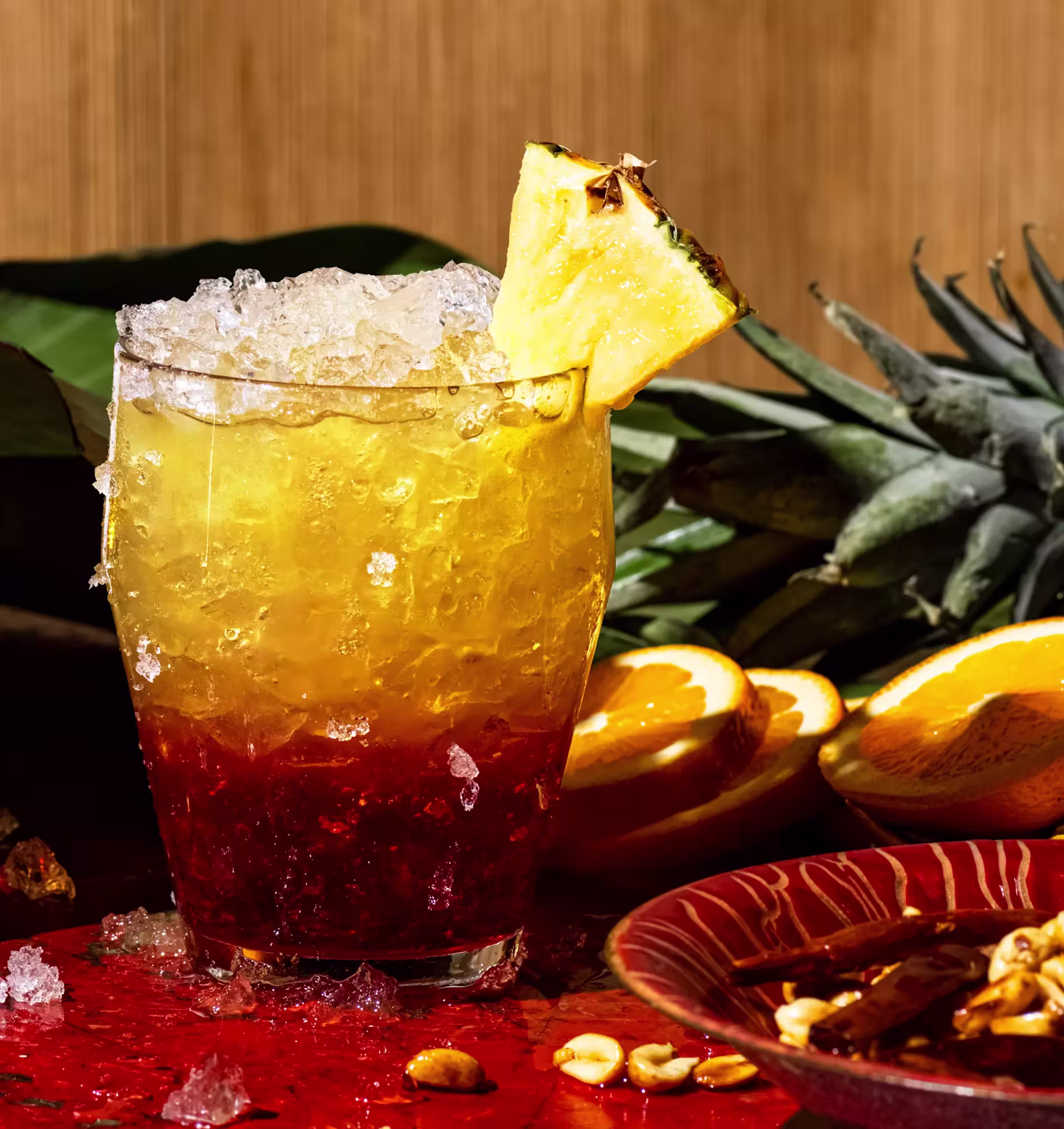 A glass filled with a layered yellow and red iced drink, garnished with a pineapple slice, sits on a table with pineapple, orange slices, and nuts nearby.