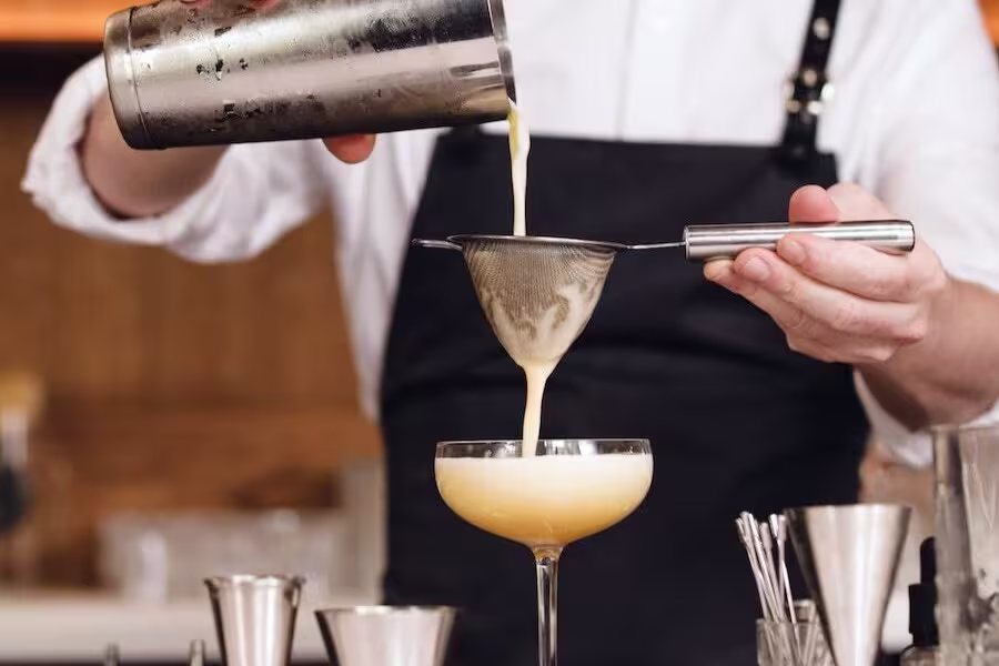 Bartender straining a creamy cocktail from a shaker into a coupe glass using a fine mesh strainer.