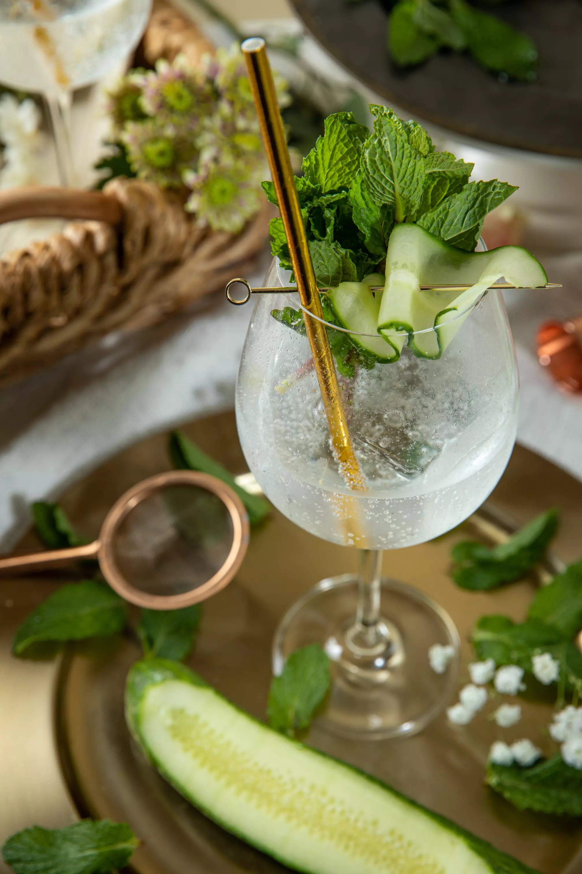 A gin and tonic with ice, cucumber ribbons, and mint garnish in a glass, placed on a gold tray with cucumber slice and mint leaves nearby.