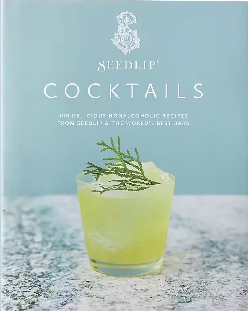 A glass of yellow-green nonalcoholic cocktail with ice and a garnish sits in front of a Seedlip Cocktails recipe book cover.