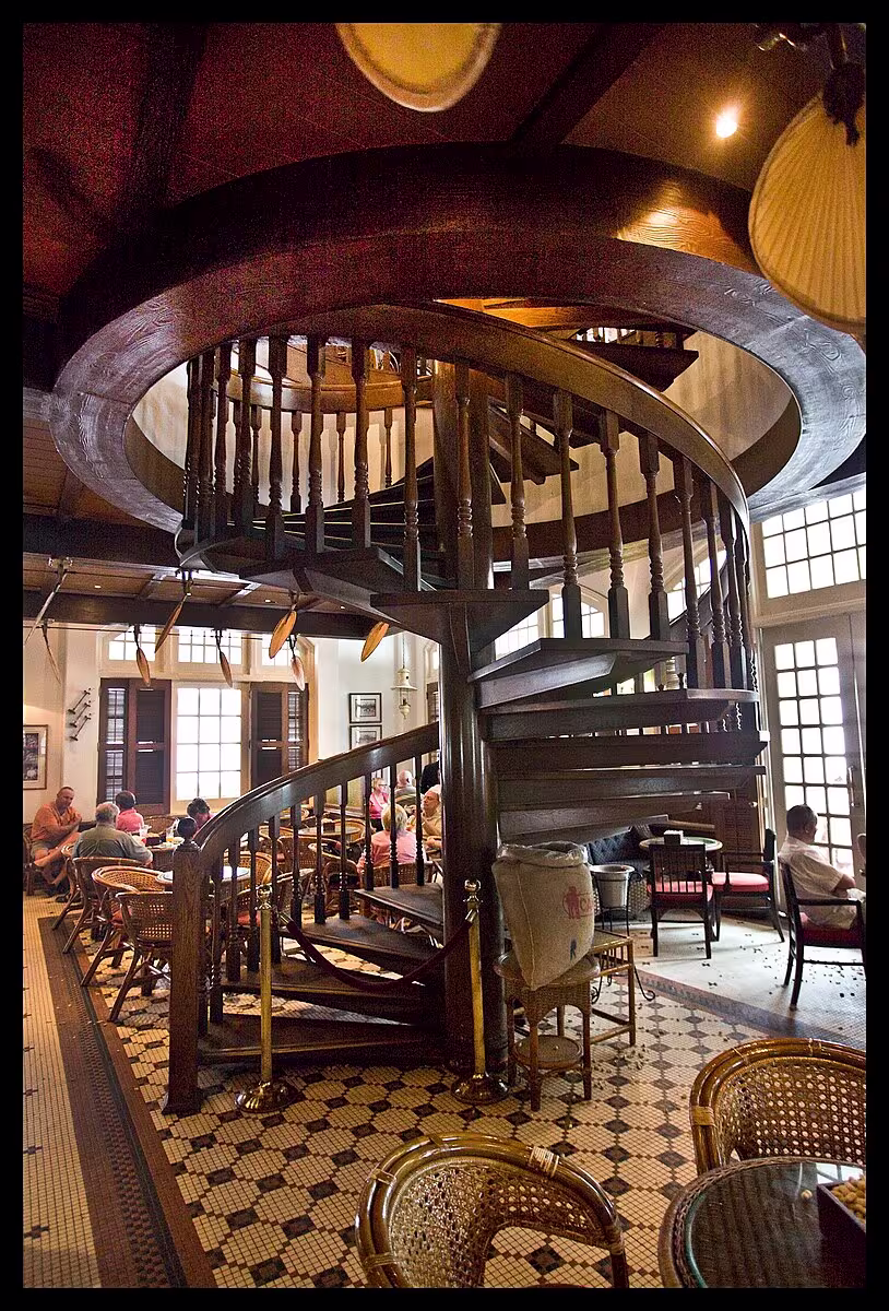 A wooden spiral staircase stands in the center of a café with patterned tile flooring, wicker chairs, and large windows letting in natural light.