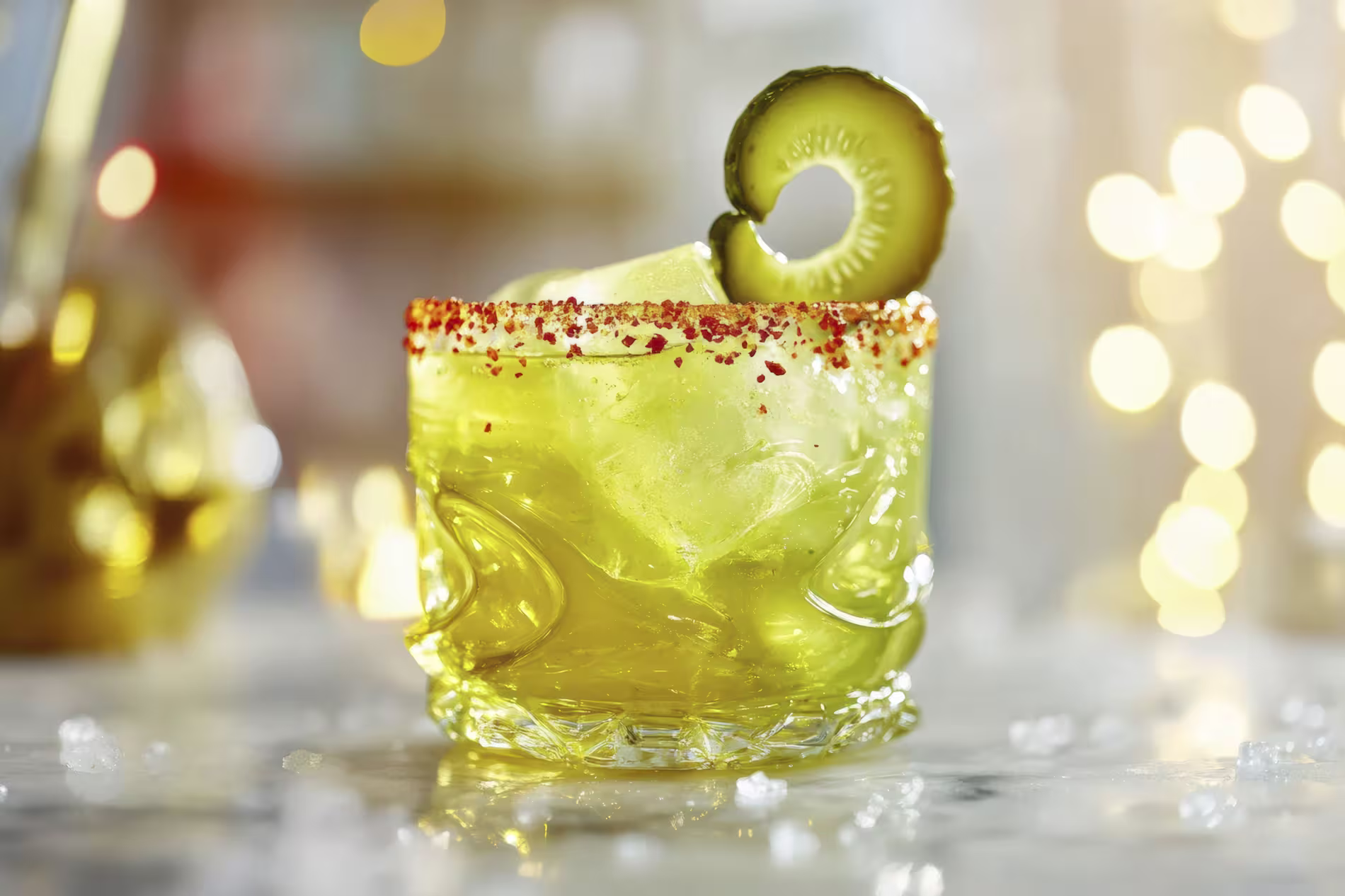 A yellow-green cocktail with ice in a clear glass, rimmed with chili powder and garnished with a pickle slice.