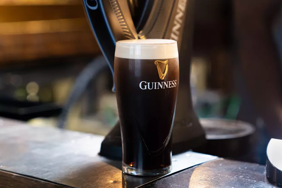 A pint glass of Guinness stout with a creamy head sits on a bar counter, with a beer tap in the background.