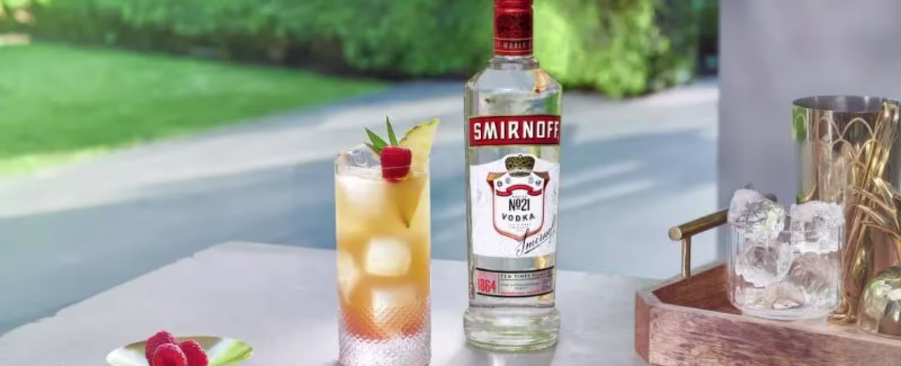 La imagen muestra una botella de vodka Smirnoff n°21 junto a un vaso relleno con un cóctel preparado con dicho destilado y otros elementos de coctelería alrededor.