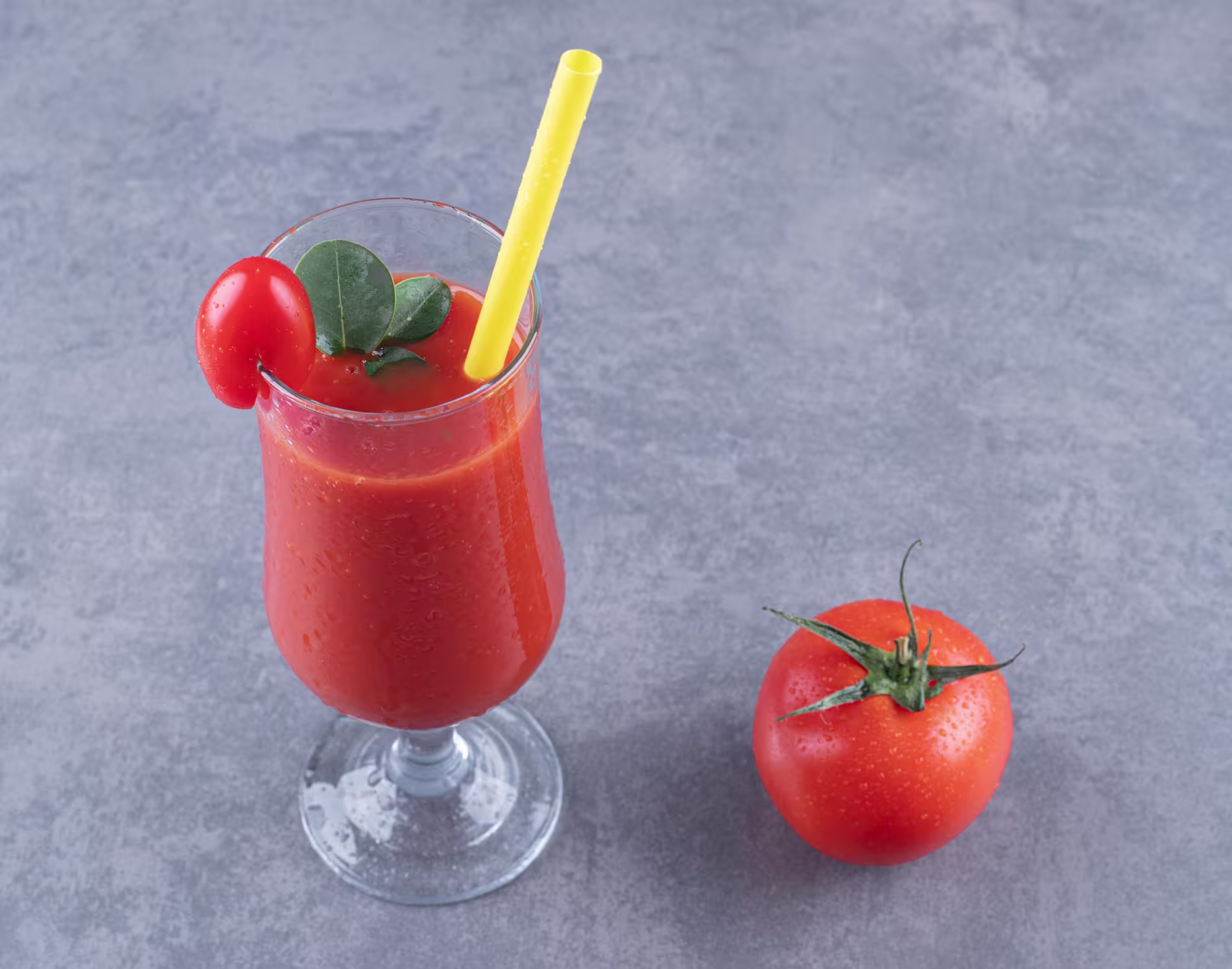 A glass of tomato juice with a yellow straw and garnish sits next to a whole tomato on a gray surface.
