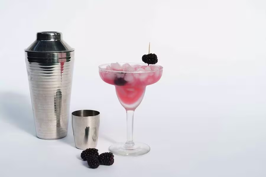 A metal cocktail shaker and jigger are next to a margarita glass filled with a pink, iced drink, garnished with blackberries.