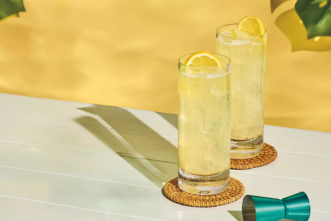 Two tall glasses of a light yellow drink with ice and lemon slices sit on woven coasters on a white table, with a green jigger nearby and a yellow background.