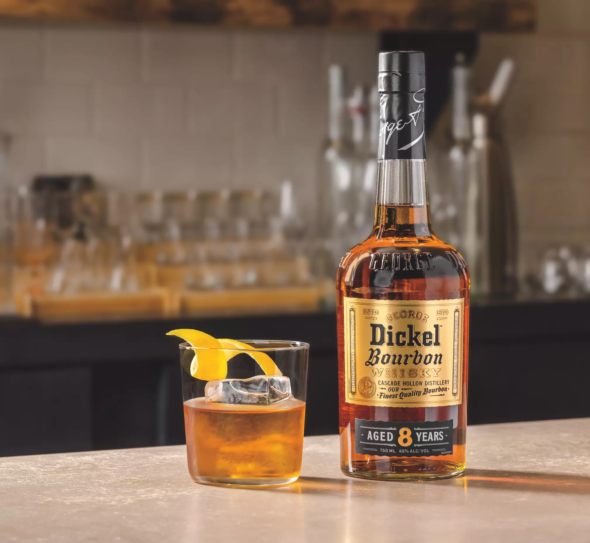 A bottle of George Dickel bourbon whiskey labeled Aged 8 Years stands next to a glass of whiskey with ice and a lemon twist on a bar counter.