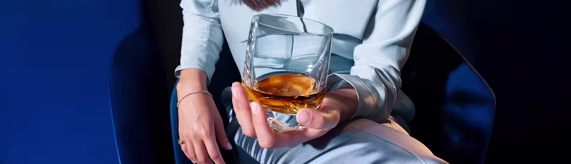 A person in light blue clothing sits on a dark chair, holding a glass of whiskey with ice, viewed from above against a dark blue background.