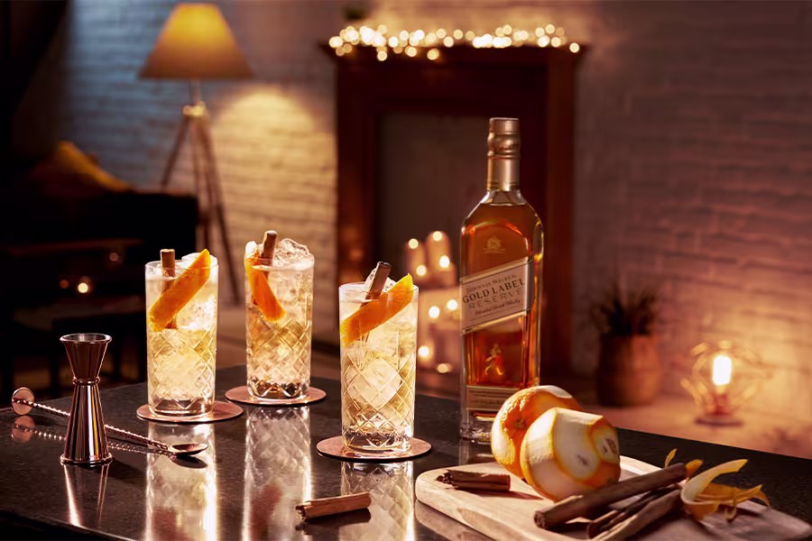 Three cocktails garnished with orange peel and cinnamon sticks are arranged beside a bottle of Johnnie Walker Gold Label, bar tools, orange peels, and cinnamon sticks in a warmly lit room.
