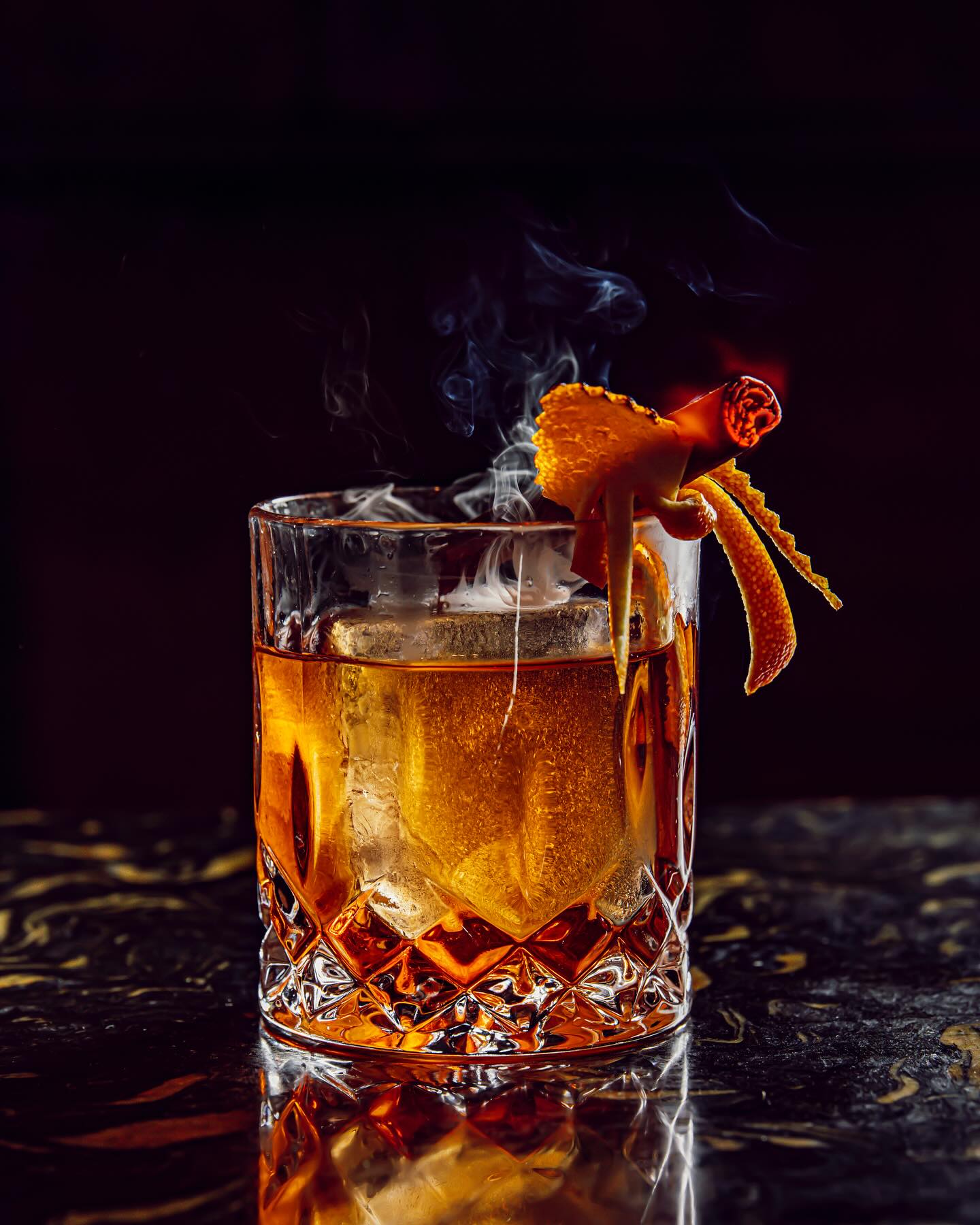 En Gangtas, un cóctel ámbar servido en un vaso tallado se presenta con una guarnición de canela ahumada y piel de naranja, resaltando su elegancia.